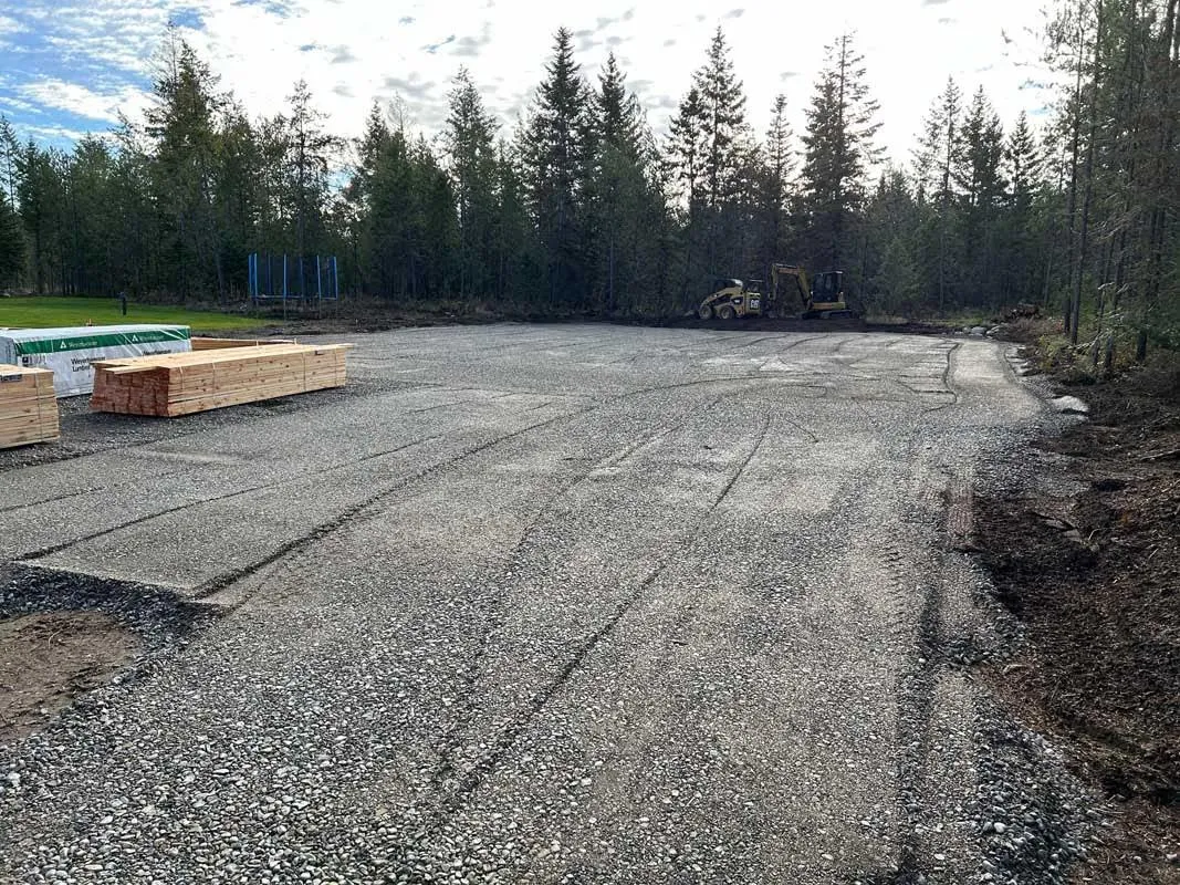 Freshly graded gravel pad surrounded by evergreen trees with an excavator in the background